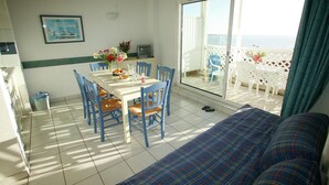 A room with a dining table, chairs, a sofa, and a view of the sea through large windows. - Apartment in France with Ocean Views (Fouras)
