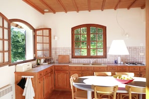 Cozinha com armários de madeira, bancada branca, janela com vista para vegetação e mesa com cadeiras.