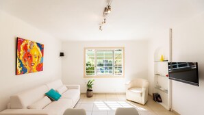 Un salón moderno con un sofá color beige, un sillón blanco, una mesa de centro de vidrio, un televisor de pantalla plana, un cuadro colorido y una ventana con vistas a la vegetación.