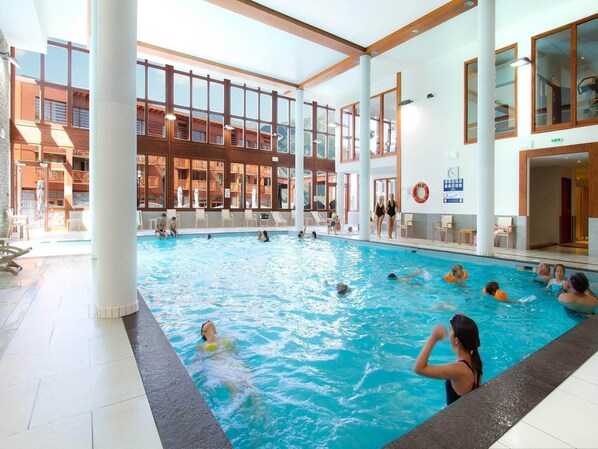An indoor swimming pool with multiple lanes, surrounded by a large glass wall, and a wooden deck area with lounge chairs.