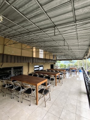 A covered outdoor dining area with tables and chairs, a grill, and a view of greenery. - Hotel Nirwana Batu (Batu)