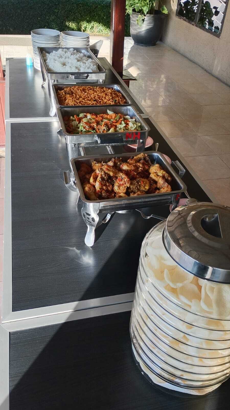 A buffet setup with multiple food trays, a stack of plates, and a large container of rice.
