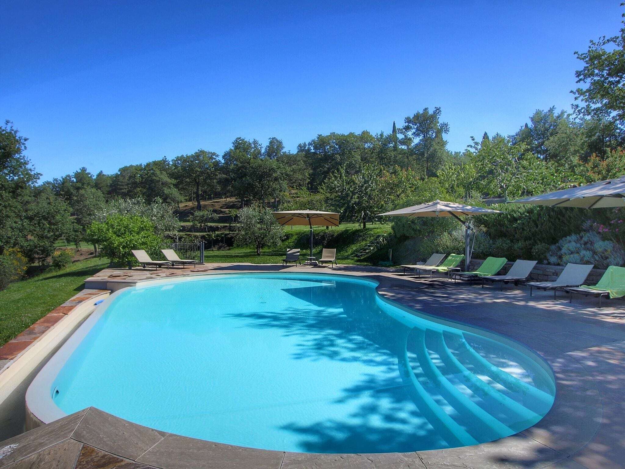 Una piscina de vora corbada, envoltada de gandules i para-sols, amb fons de paisatge de bosc i cel blau.