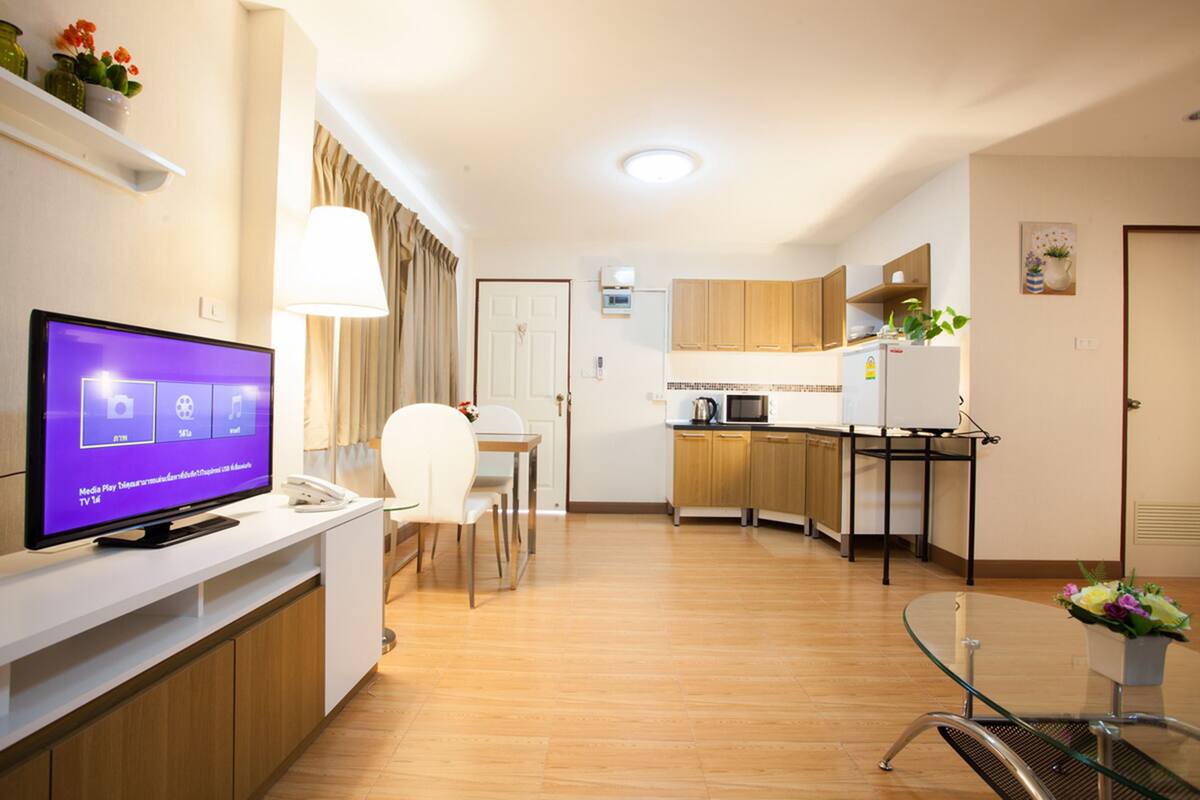 A modern hotel room with a TV, a dining area, a kitchen, and a glass table.