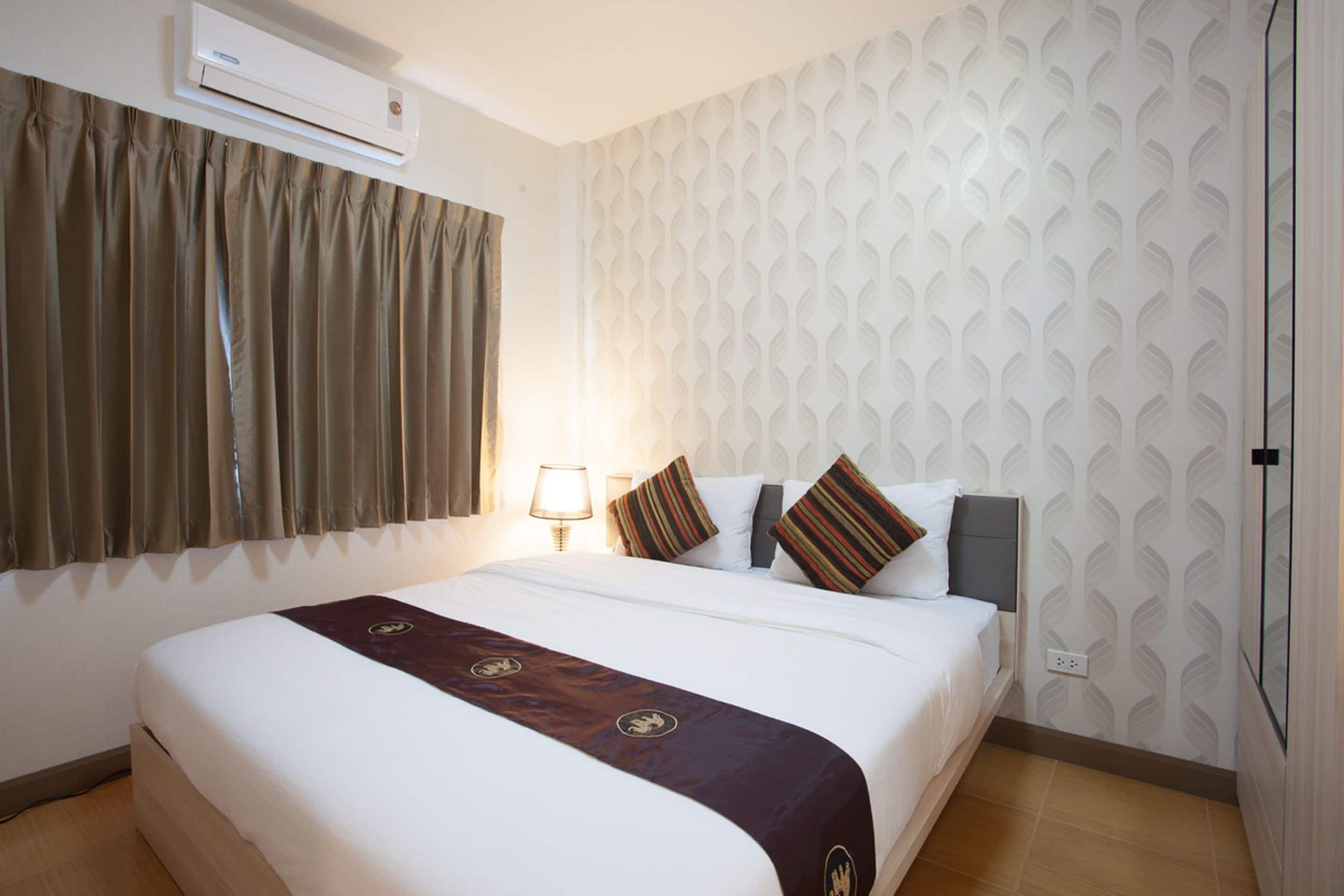 A hotel room with a large bed, a wall-mounted air conditioner, a window with curtains, and a patterned wallpaper.