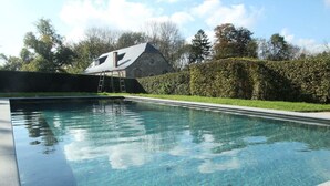 Una piscina limpia con una escalera de acceso, rodeada de un seto y un edificio con techo inclinado al fondo.
