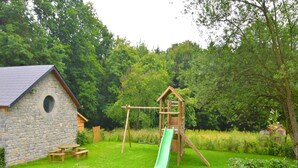 Un edificio de piedra con una ventana circular, una estructura de madera con un tobogán y una mesa de picnic en un área cubierta de césped con árboles al fondo.