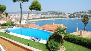 A swimming pool with lounge chairs, a palm tree, and a view of a coastal cityscape.