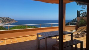 A balcony with a table and chairs, overlooking a coastal view with a clear sky.