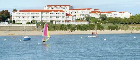 A coastal hotel with multiple balconies, a beach in the background, and a sailboat on the water.