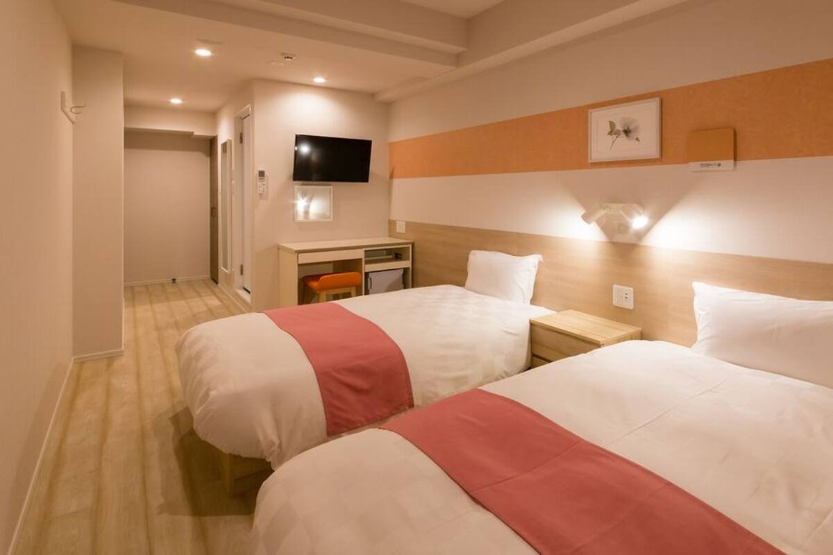 A hotel room with two beds, a desk, a television, and a small kitchenette.