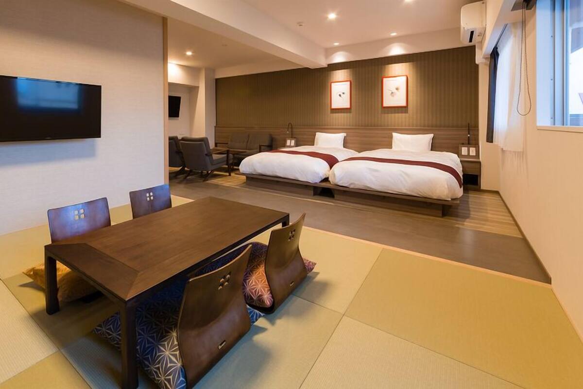 A hotel room with a wooden dining table, two beds, a flat-screen TV, and a large window.