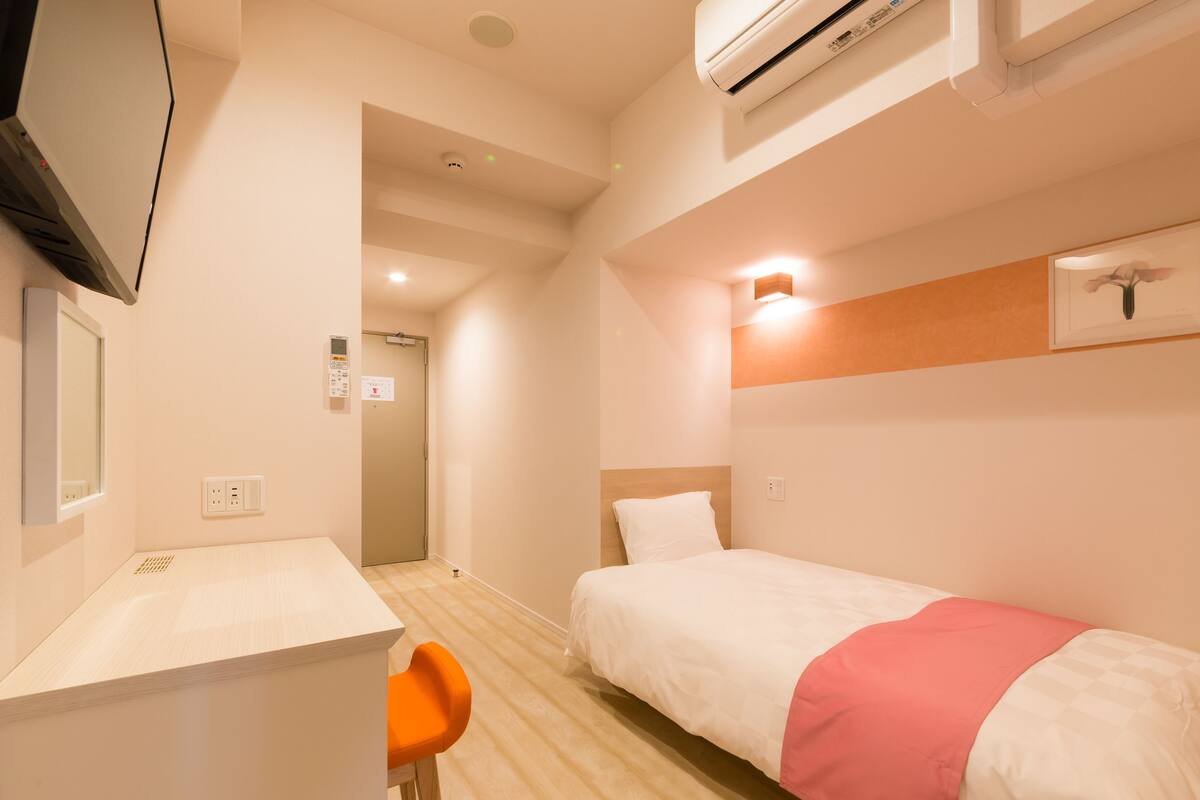 A hotel room with a bed, a desk, and a chair. There is an air conditioning unit on the wall and a door leading to another room.