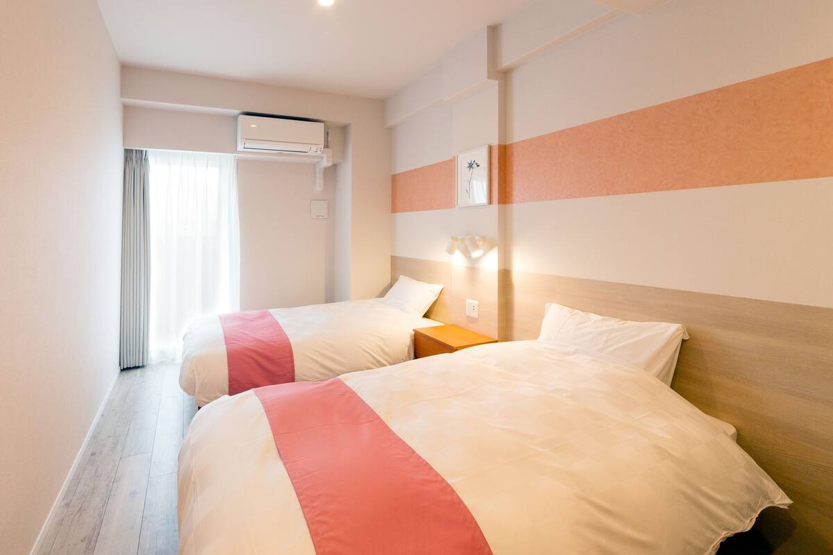 A hotel room with two beds, a window with curtains, an air conditioning unit, and a wall-mounted light fixture.