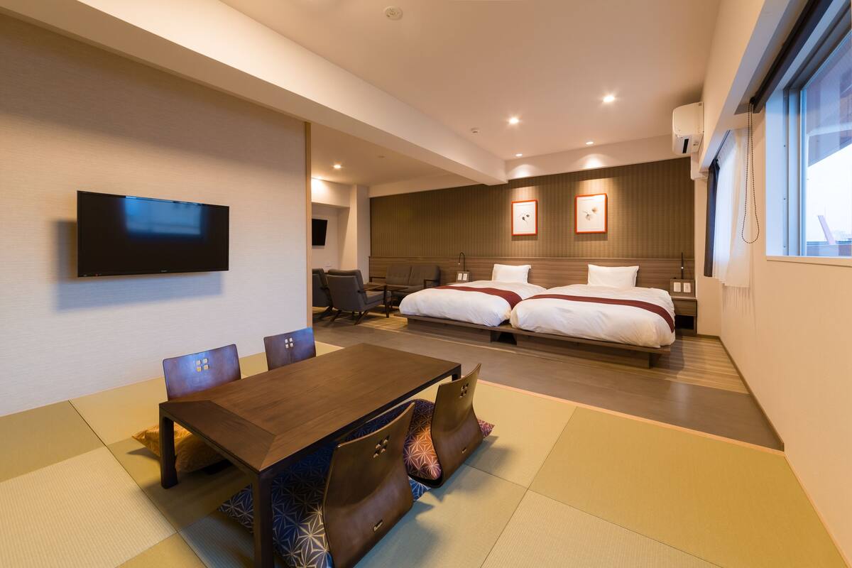 A hotel room with a wooden dining table, two beds, a TV, and a large window.