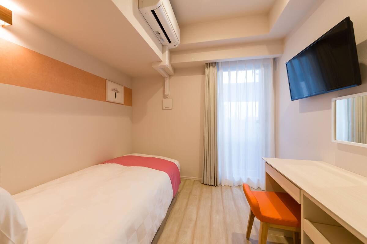 A hotel room with a bed, a desk, a chair, an air conditioning unit, a television, and a window with curtains.
