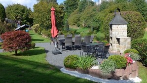 Un'area patio con posti a sedere all'aperto, una zona barbecue in pietra e un piccolo giardino con diverse piante e un ombrello rosso.