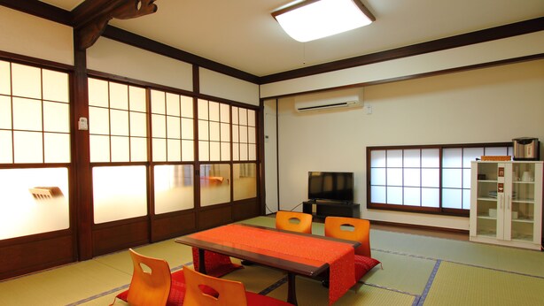 Una habitación japonesa tradicional con suelo de tatami, una mesa baja con mantel rojo, sillas anaranjadas y un televisor.