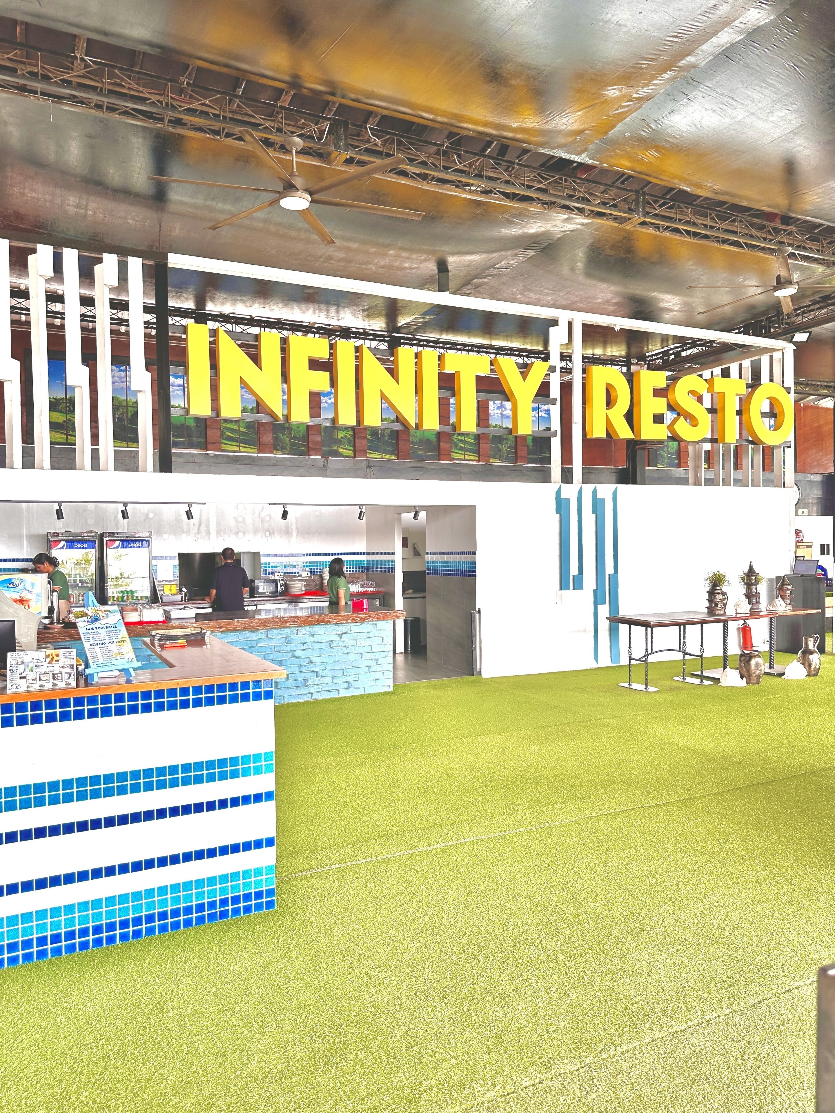 En restauranginteriör med en skylt som visar texten ”INFINITY RESTO” och en bardisk med blå kakelplattor på väggen bakom och ett ställ med menyer.