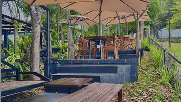 Relax under the shade of large umbrellas in a serene outdoor seating area surrounded by lush greenery and a natural fence.
