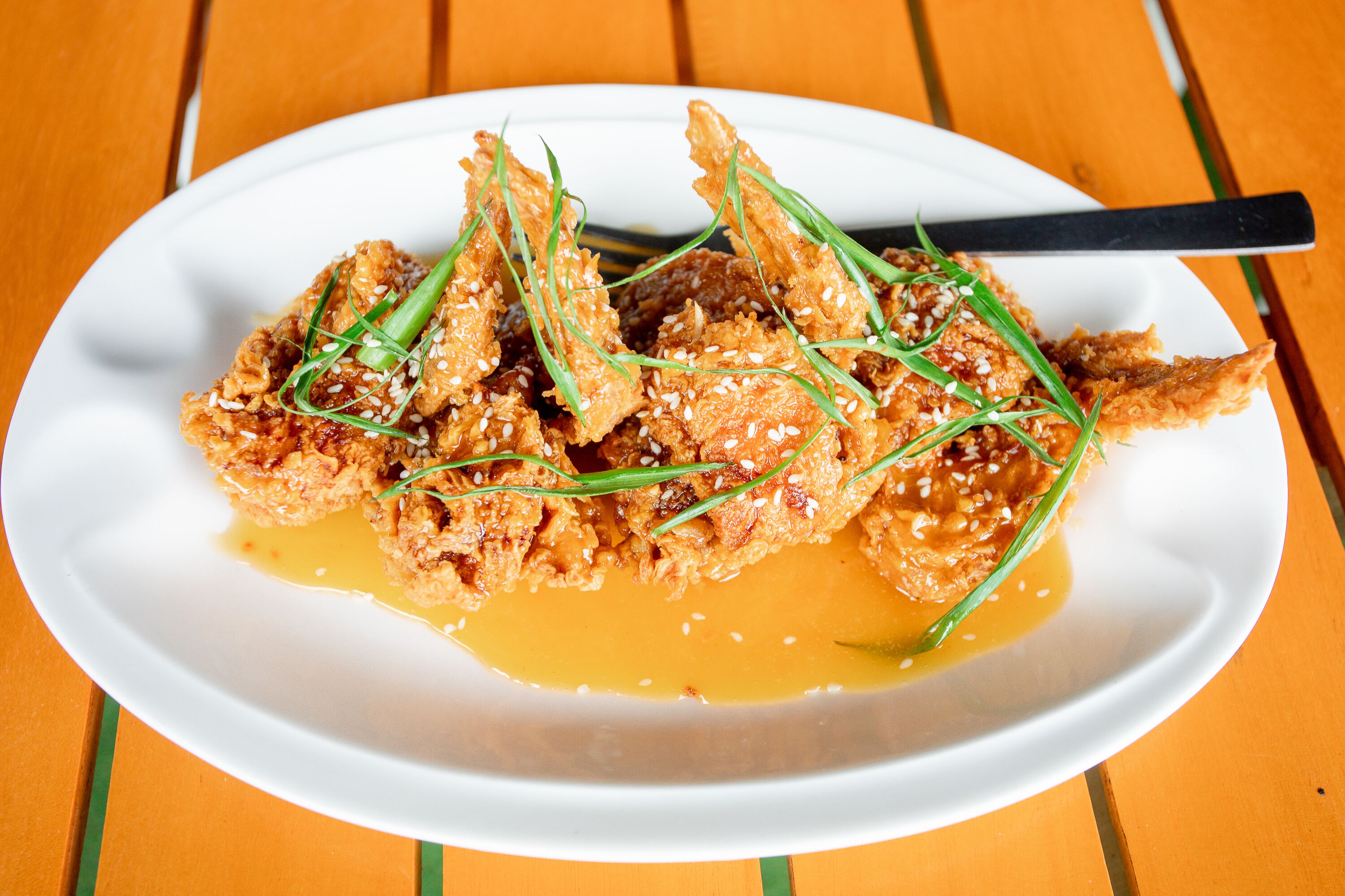 A white plate with fried chicken pieces, garnished with green herbs and sesame seeds, on a wooden table.