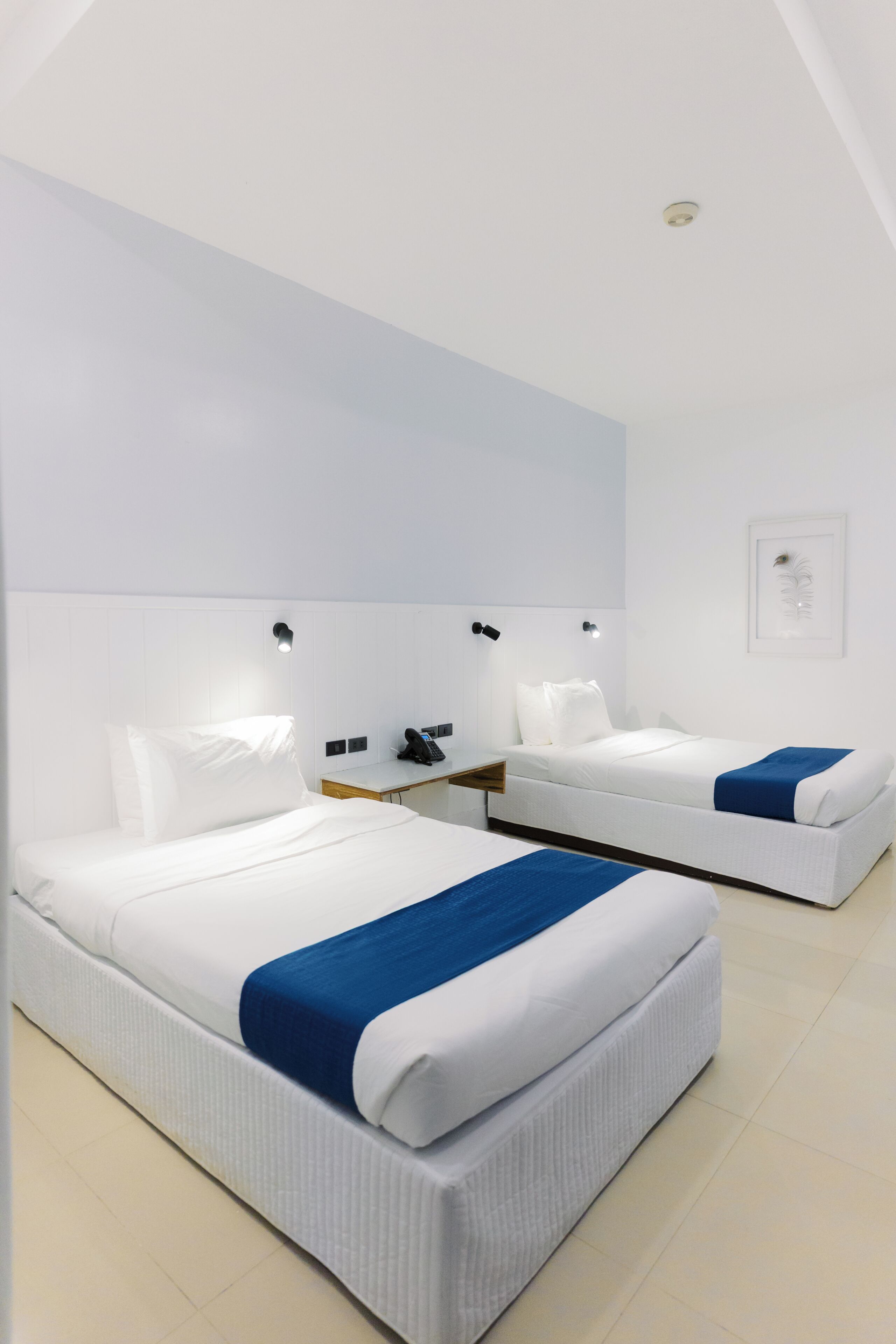 A hotel room with two beds, a bedside table with a phone, and a wall-mounted light fixture.