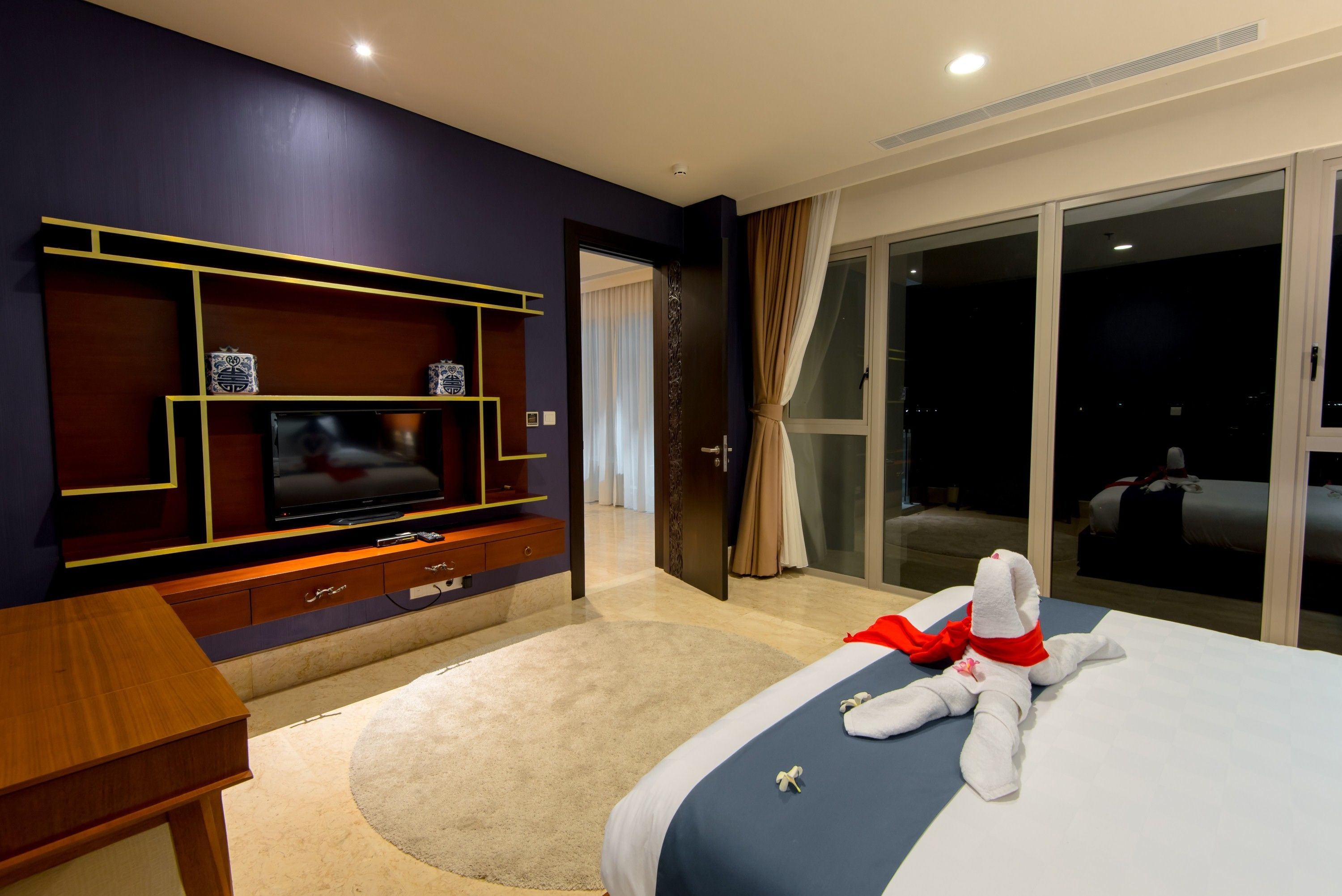 A hotel room with a large bed, a television on a wooden stand, a desk, and a view of the outside through a sliding glass door.