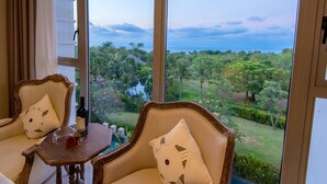 Ein Zimmer mit zwei Stühlen und einem kleinen Tisch, jeweils mit einem kissenförmigen Katzenkissen. Auf dem Tisch steht eine Weinflasche und ein Glas. Das Zimmer verfügt über große Fenster mit Blick auf einen üppigen Garten und ein Gewässer.