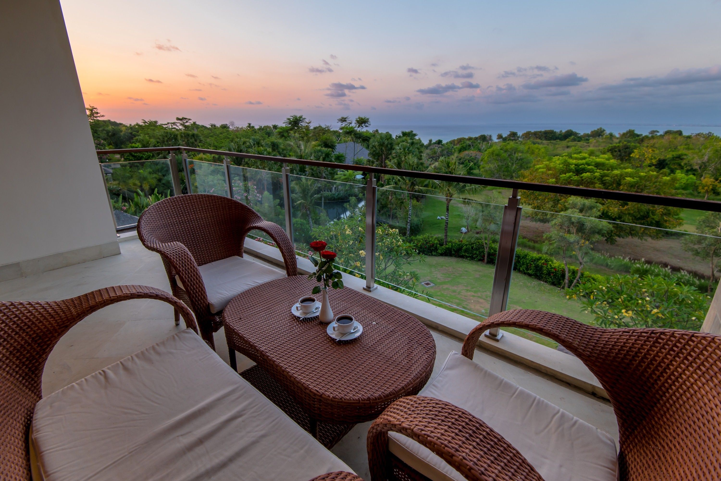Un balcon agrémenté de meubles en osier, une table avec des tasses et un vase, et une vue sur la verdure et la mer.