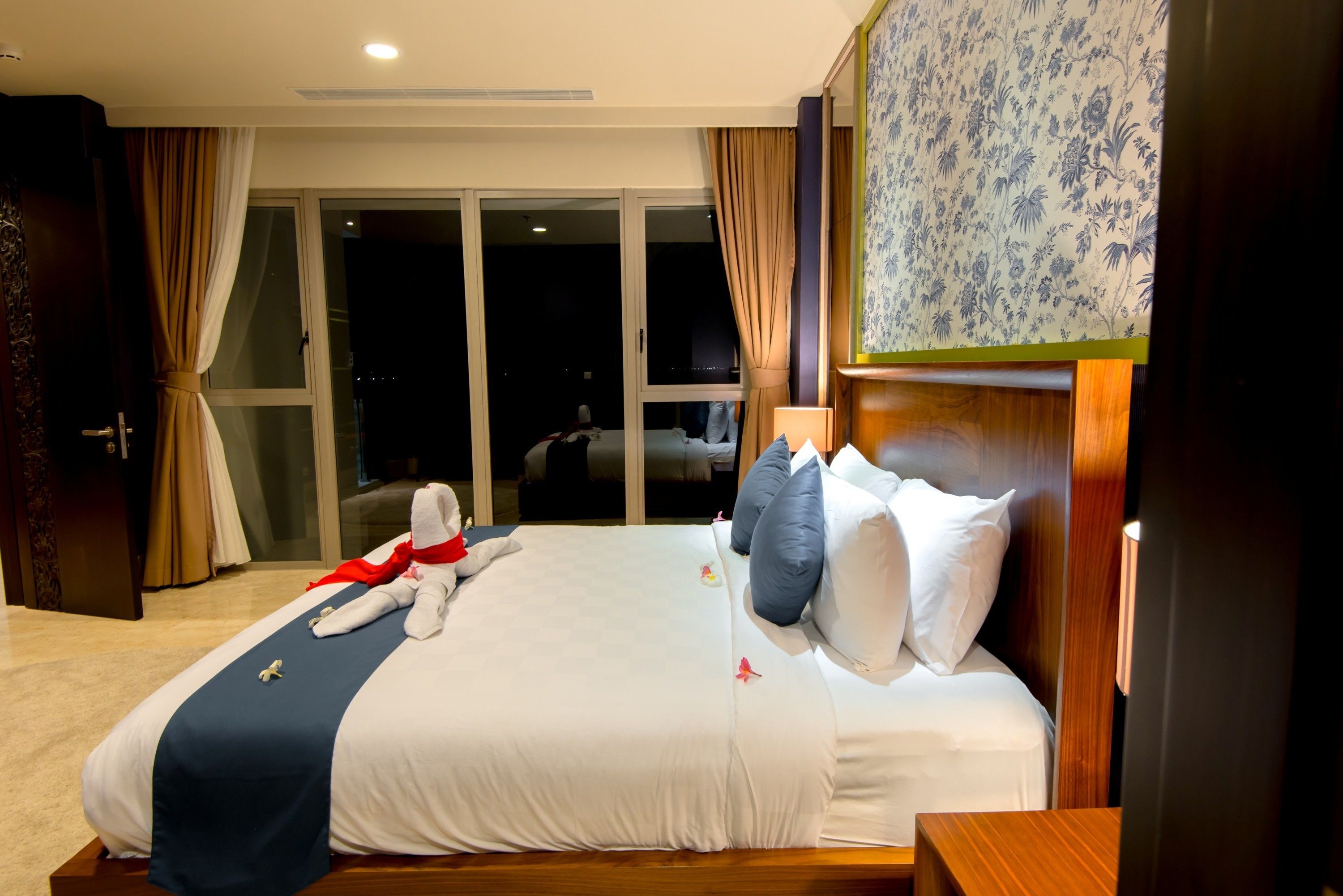 A hotel room with a large bed, a wooden headboard, a nightstand, and a view of the outside through a sliding glass door.