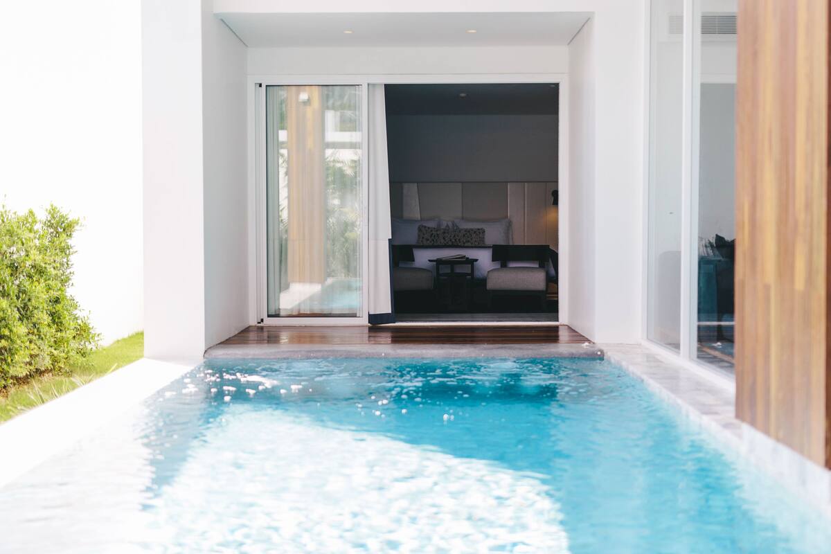 Una piscina interior moderna con vistas despejadas a un dormitorio, que incluye una cama, mesitas de noche y un ventanal.