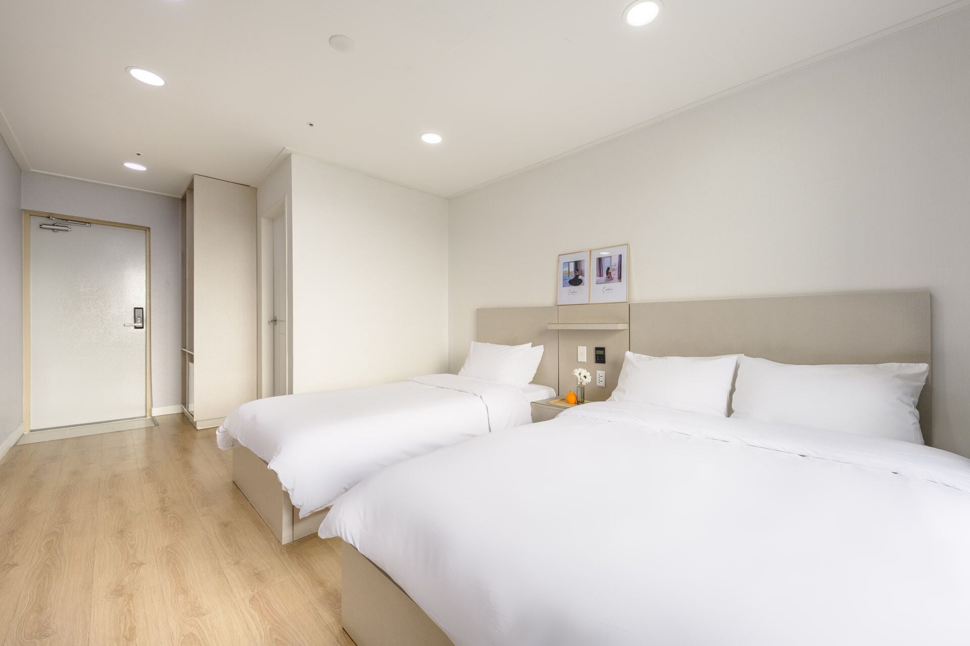 Ein Hotelzimmer mit zwei Einzelbetten, einem Nachttisch mit einer Vase, einer Wandleuchte und einer Tür mit Griff.