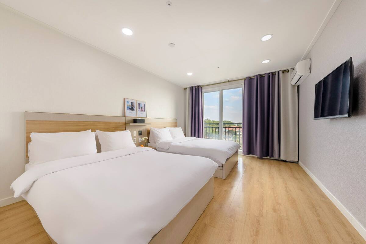 Une chambre d’hôtel avec deux lits, une grande fenêtre avec des rideaux, un téléviseur fixé au mur et un plafond doté d’un éclairage encastré.