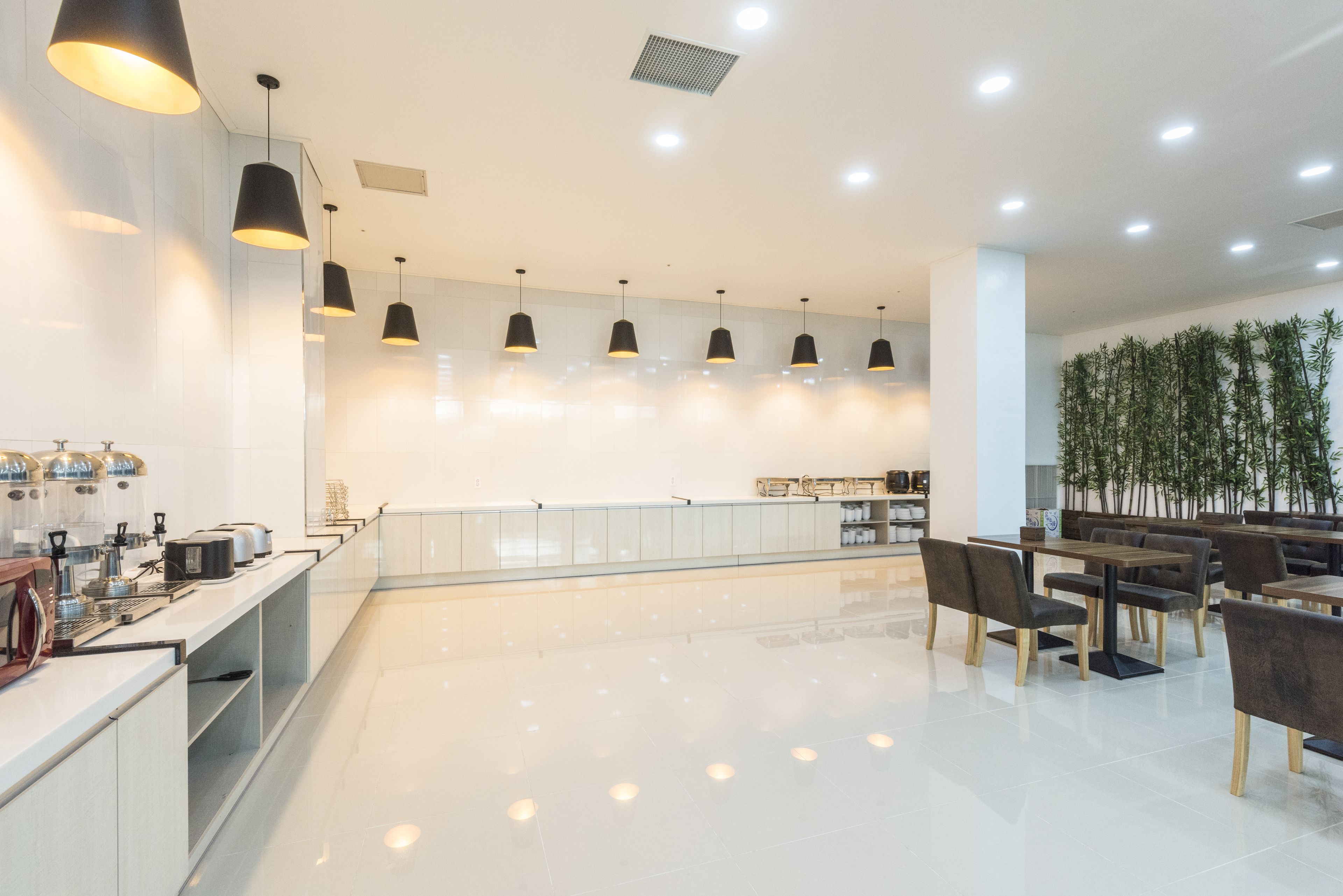 A modern cafe with a long counter, multiple hanging pendant lights, and a seating area with tables and chairs.