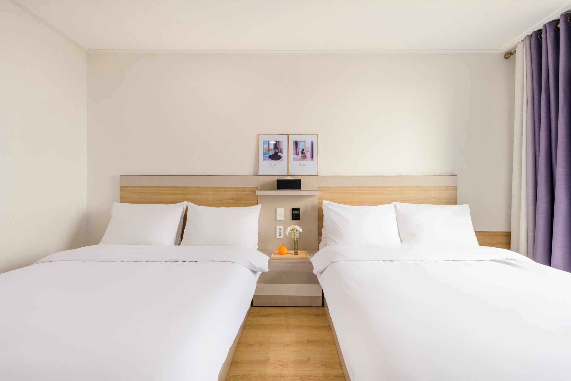 A hotel room with two beds, a wooden headboard, a nightstand with a radio, and a small table with a vase.