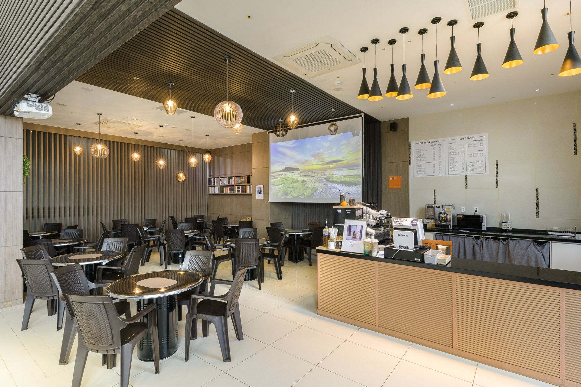 A modern cafe with multiple seating areas, a counter with a coffee machine, and a large screen displaying a scenic view.