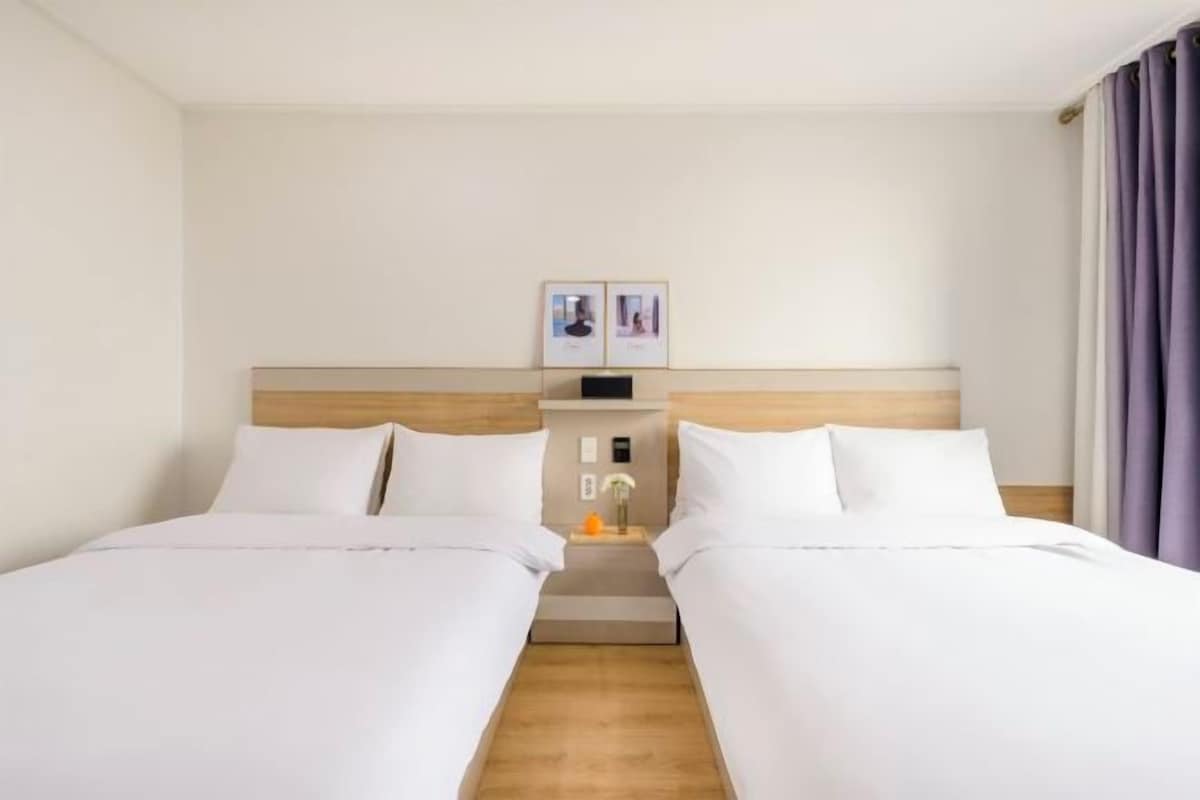 Ein Hotelzimmer mit zwei Betten, einem hölzernen Kopfteil, einem Nachttisch mit Lampe sowie gerahmten Bildern an der Wand.