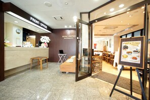 Un espace d’accueil d’hôtel comprenant un comptoir de réception, un petit coin salon et une salle à manger visible à travers la porte ouverte.
