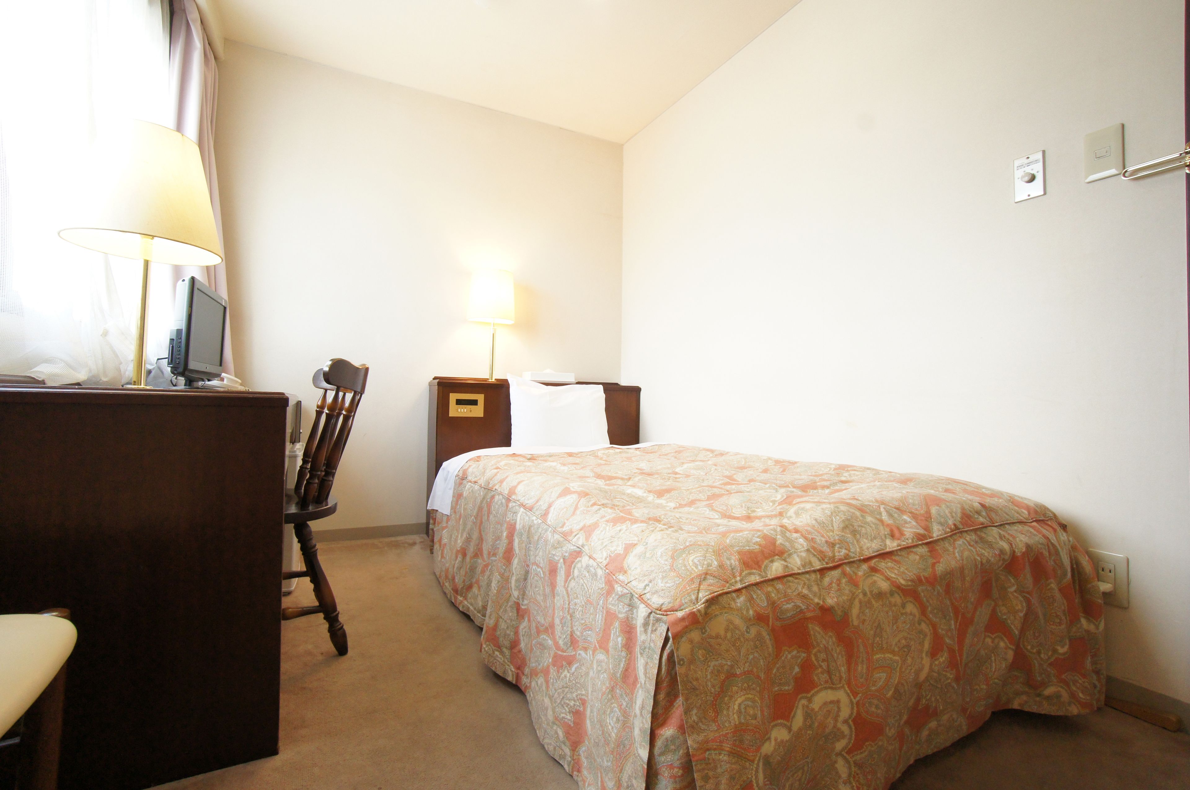 A hotel room with a bed, a desk with a lamp, a chair, and a small table.