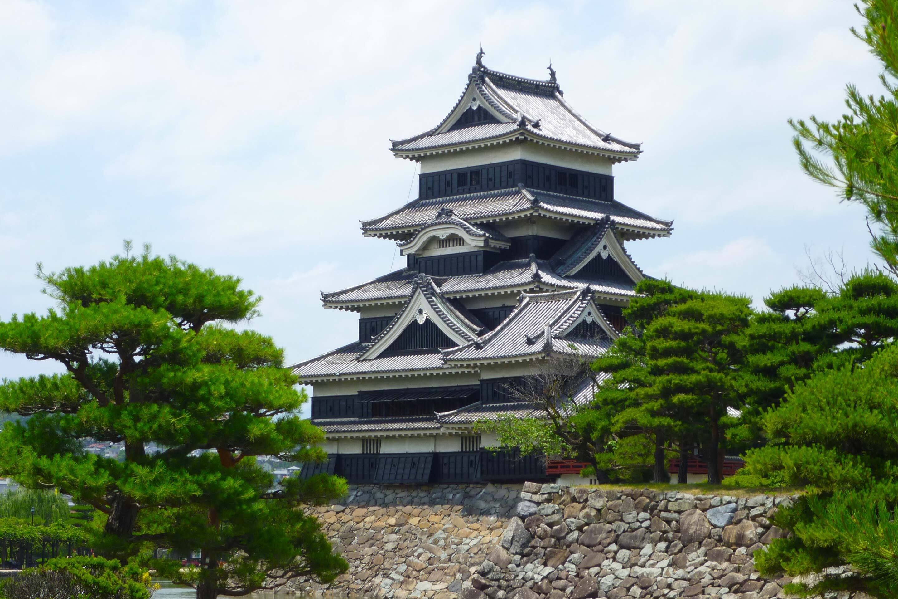 A traditional Japanese castle with multiple tiers and a stone base, surrounded by lush greenery.