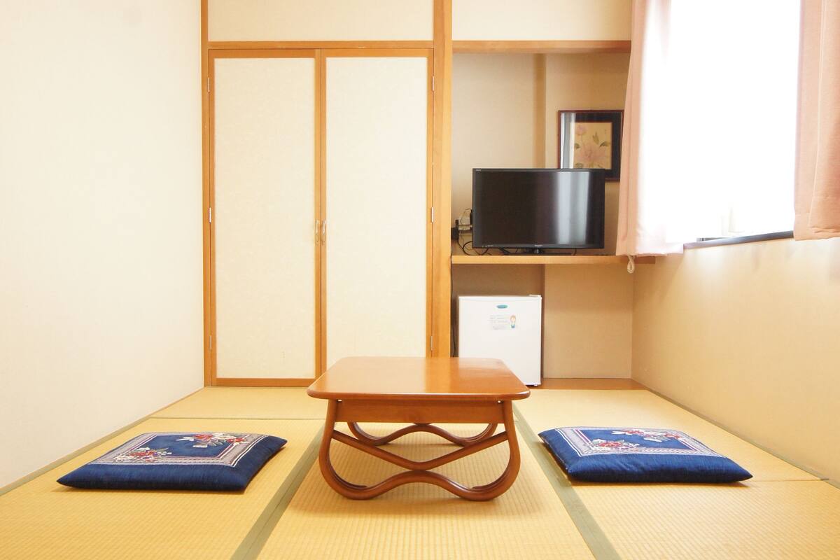 A traditional Japanese room with tatami flooring, a low wooden table, a television on a shelf, and a sliding door.