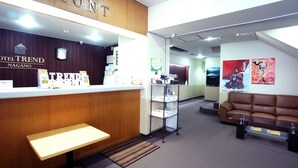 Un área de recepción con un mostrador de madera identificado con la etiqueta “FRONT”, un banco, un estante metálico con objetos y un sofá de cuero marrón.