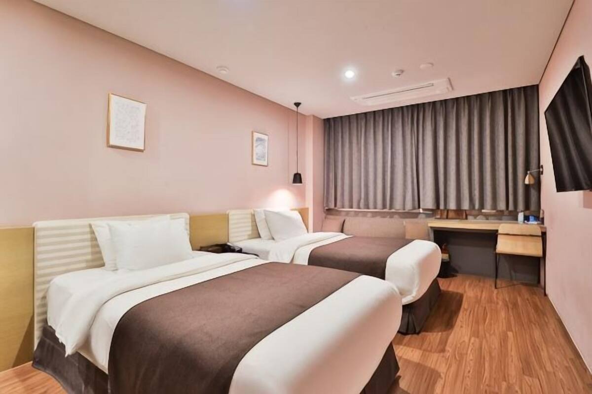 A hotel room with two beds, a desk, a chair, a window with curtains, and a wall-mounted TV.