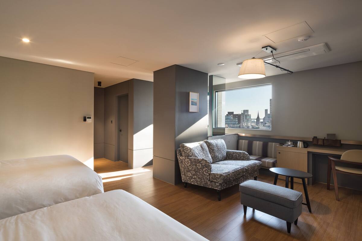 A modern hotel room with a large bed, a seating area with a patterned chair, a small table, and a city view through a large window.