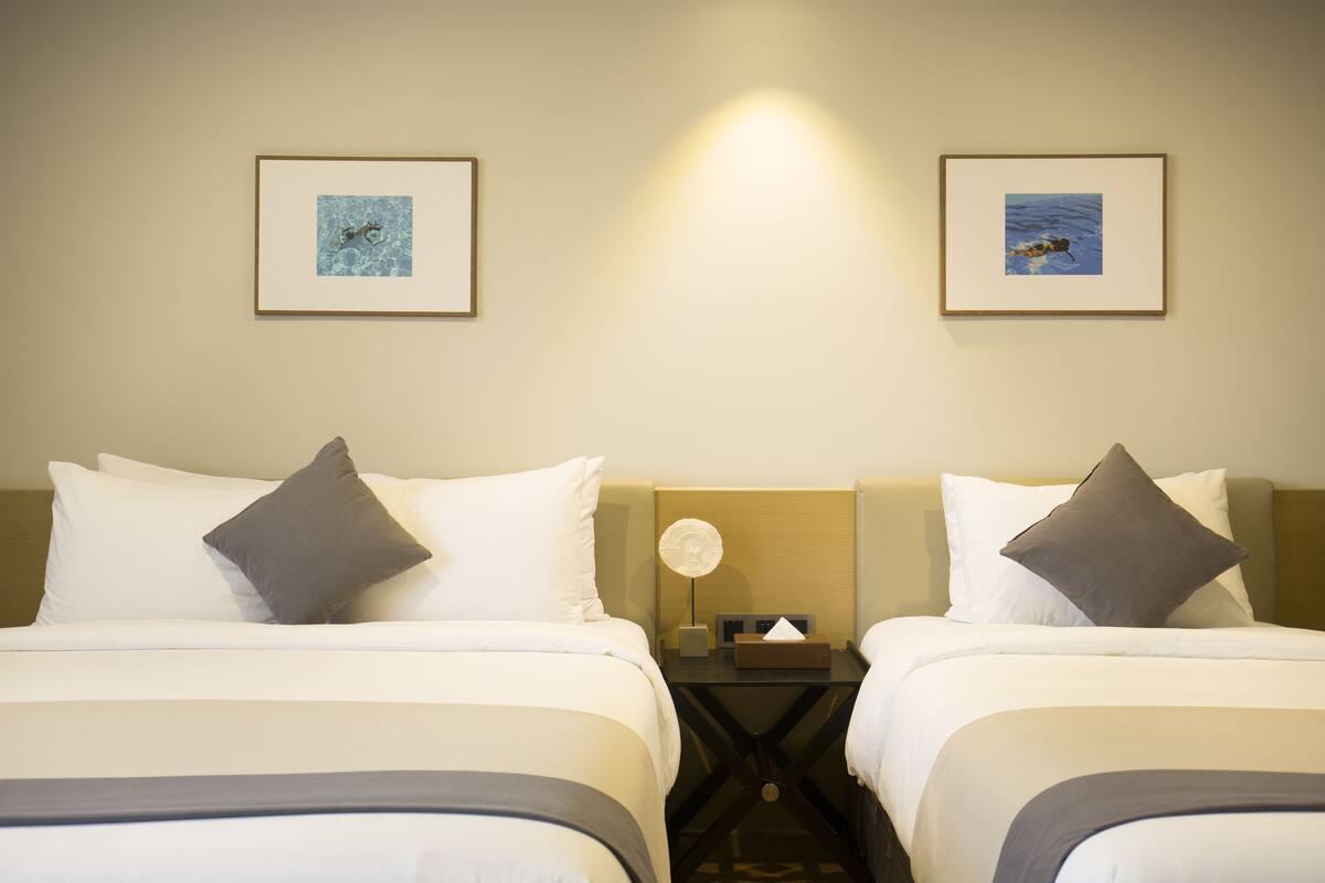 A hotel room with two beds, each with white bedding and a gray pillow. There are framed pictures on the wall, a nightstand with a lamp, and a small table with a tissue box.