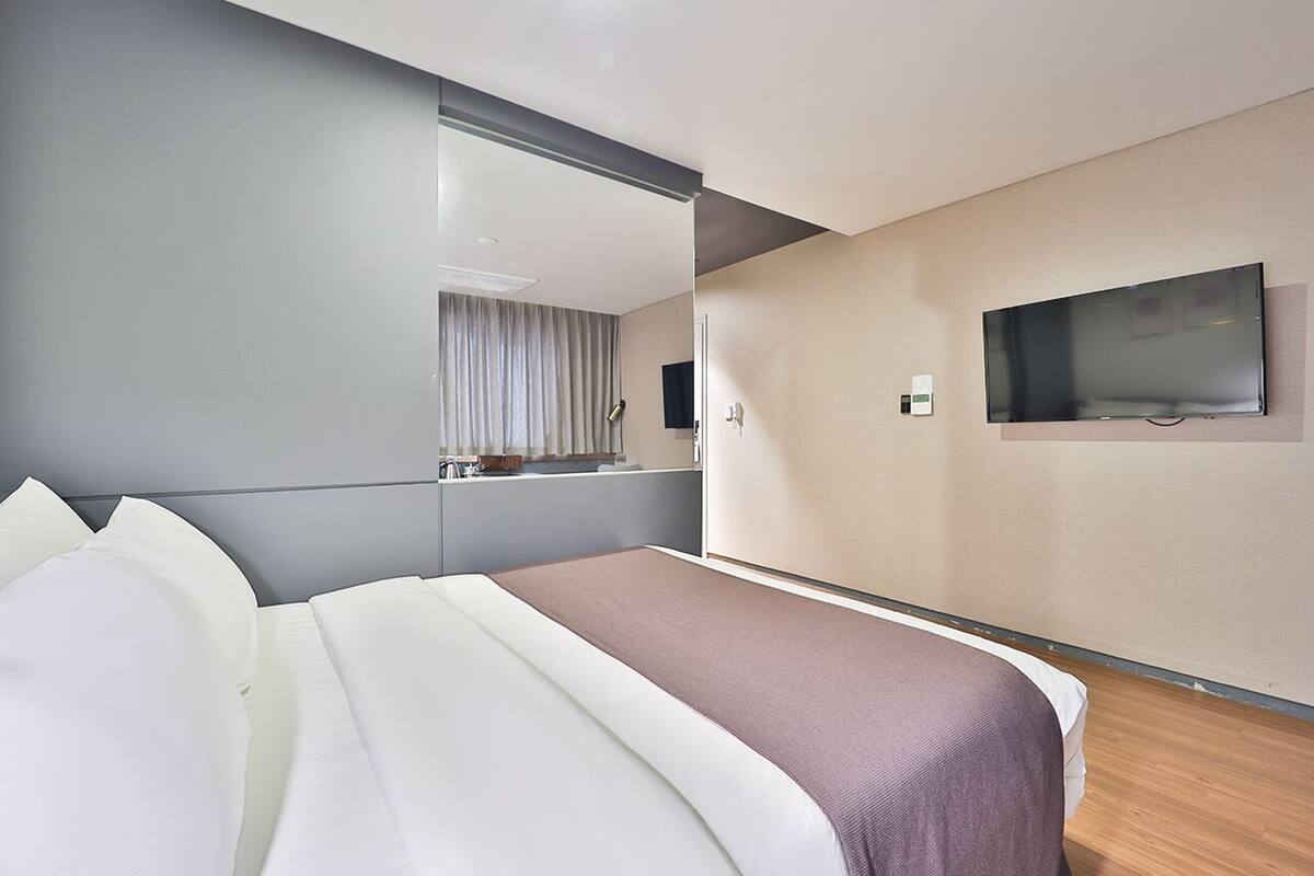 A modern hotel room with a large bed, a flat-screen TV mounted on the wall, and a mirror above the bed.