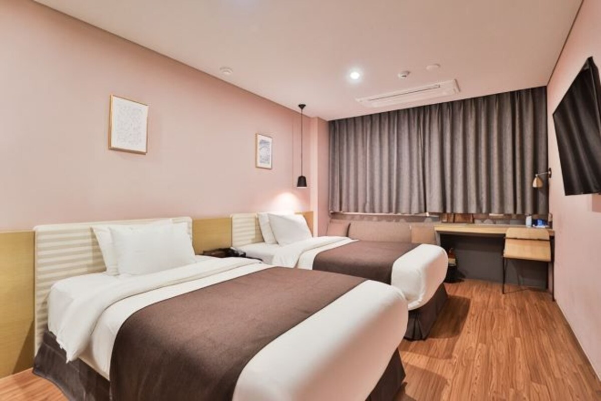 Une chambre d’hôtel avec deux lits, un bureau, une chaise, une fenêtre avec des rideaux et un téléviseur fixé au mur.