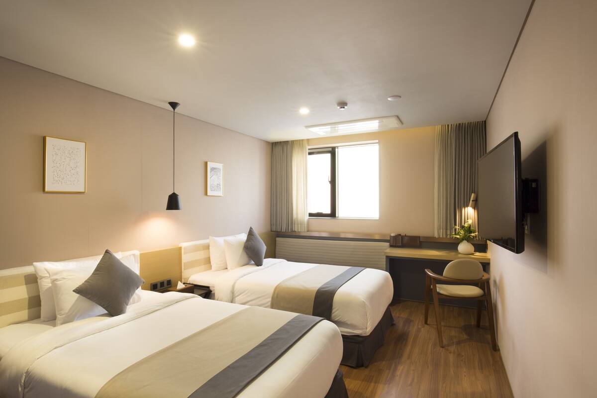 A hotel room with two beds, a desk, a chair, a TV, and a window with curtains.