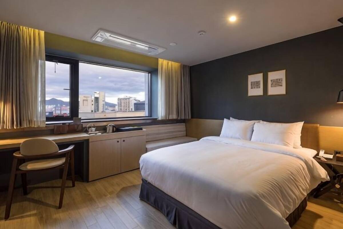 A hotel room with a bed, desk, chair, window with curtains, and a city view.