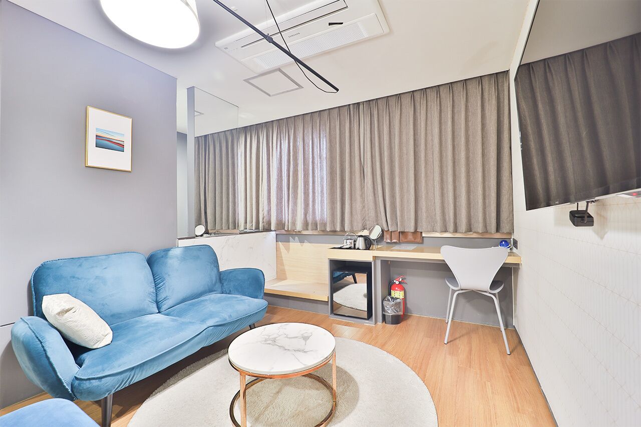 Une chambre d’hôtel moderne avec un canapé bleu, une petite table ronde, un bureau avec une chaise, une grande fenêtre avec des rideaux et un extincteur.